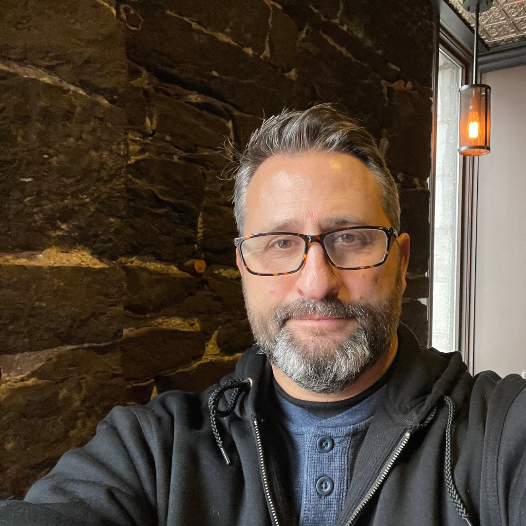 Image of Laurent Ergas, owner of Painters Pro KC, standing in front of a stone wall, wearing glasses, a black hoodie, and blue button-up shirt.